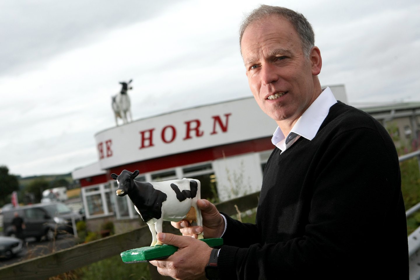 Kenny Farquharson holds a model cow outside the The Horn, with the cow on the roof visible in the background.